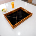 Wooden tray with black marble insert on a white surface, with a glass of orange juice and a piece of cake in the background.